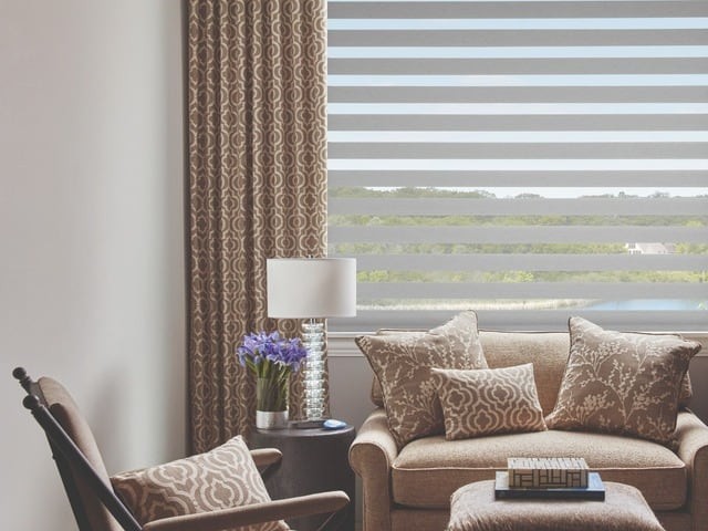 Living room featuring a large window with drapes and shades in front of accent chairs and an end table.s